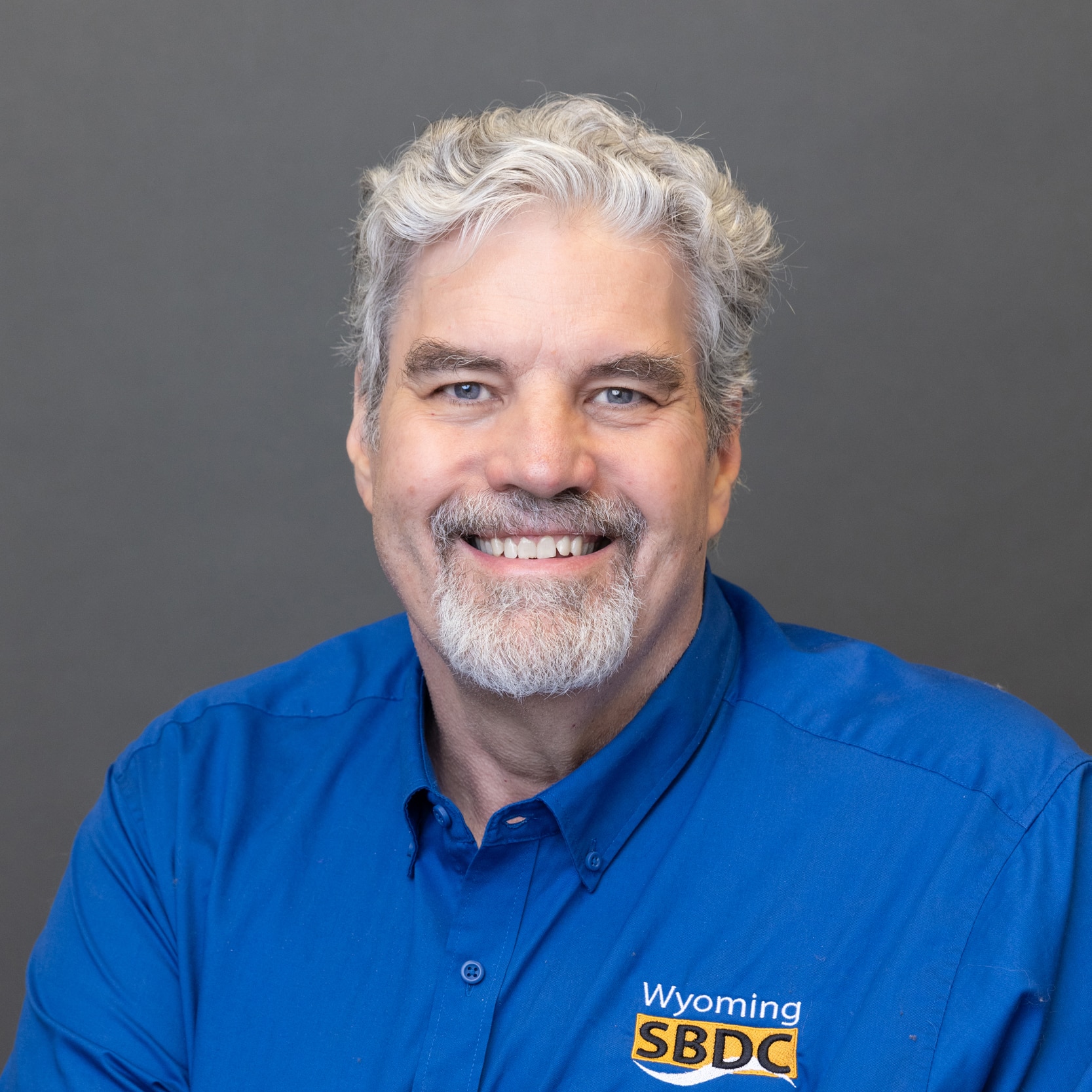 Photo of Mike Lambert in front of a plain blue background and wearing a white shirt that says "Wyoming SBDC Network"